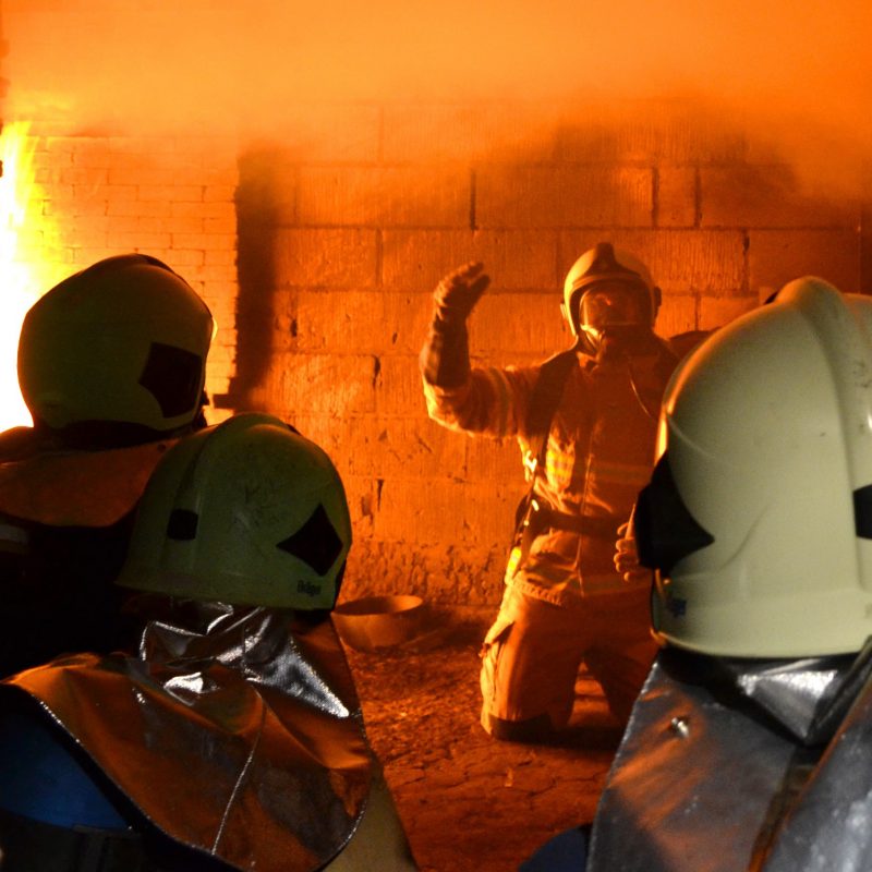 Hjælperøgdykkere under uddannelse. Fire uniformerede røddykkere sidder foran en øvelseshus i brand.